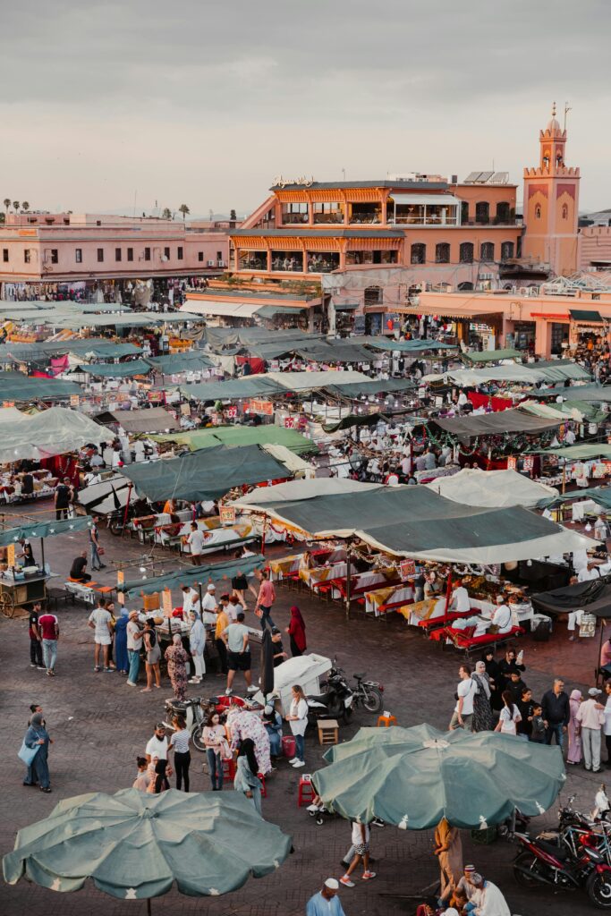 مشهد حيوي من ساحة جامع الفنا في مراكش حيث يتجمع الزوار المحليون والسياح بين أكشاك الطعام والمحلات التجارية التي تعرض الحرف اليدوية المغربية العروض الثقافية الحية مثل الموسيقى والعروض المسرحية تضفي على المكان أجواء مميزة مع أضواء المساء التي تبرز سحر الساحة