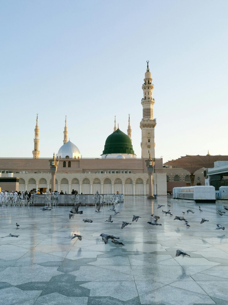 صورة لفناء المسجد النبوي في المدينة المنورة حيث يظهر المسجد بروعته والحمام يطير وسط الزوار مما يخلق أجواء من السكينة والروحانية في هذا المكان المقدس